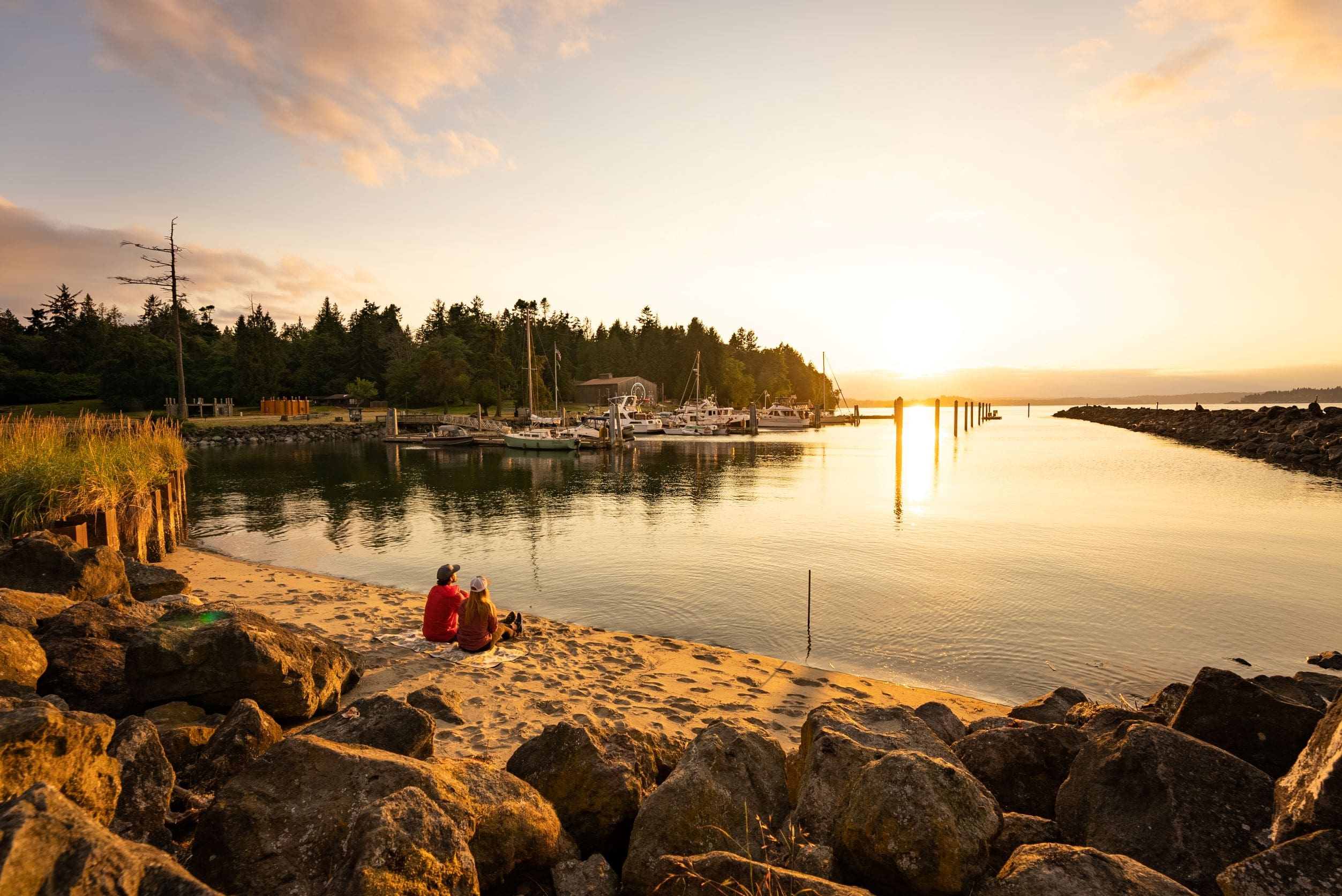 Two people sitting on beach near a boat marina with boats in midground and sunset in background