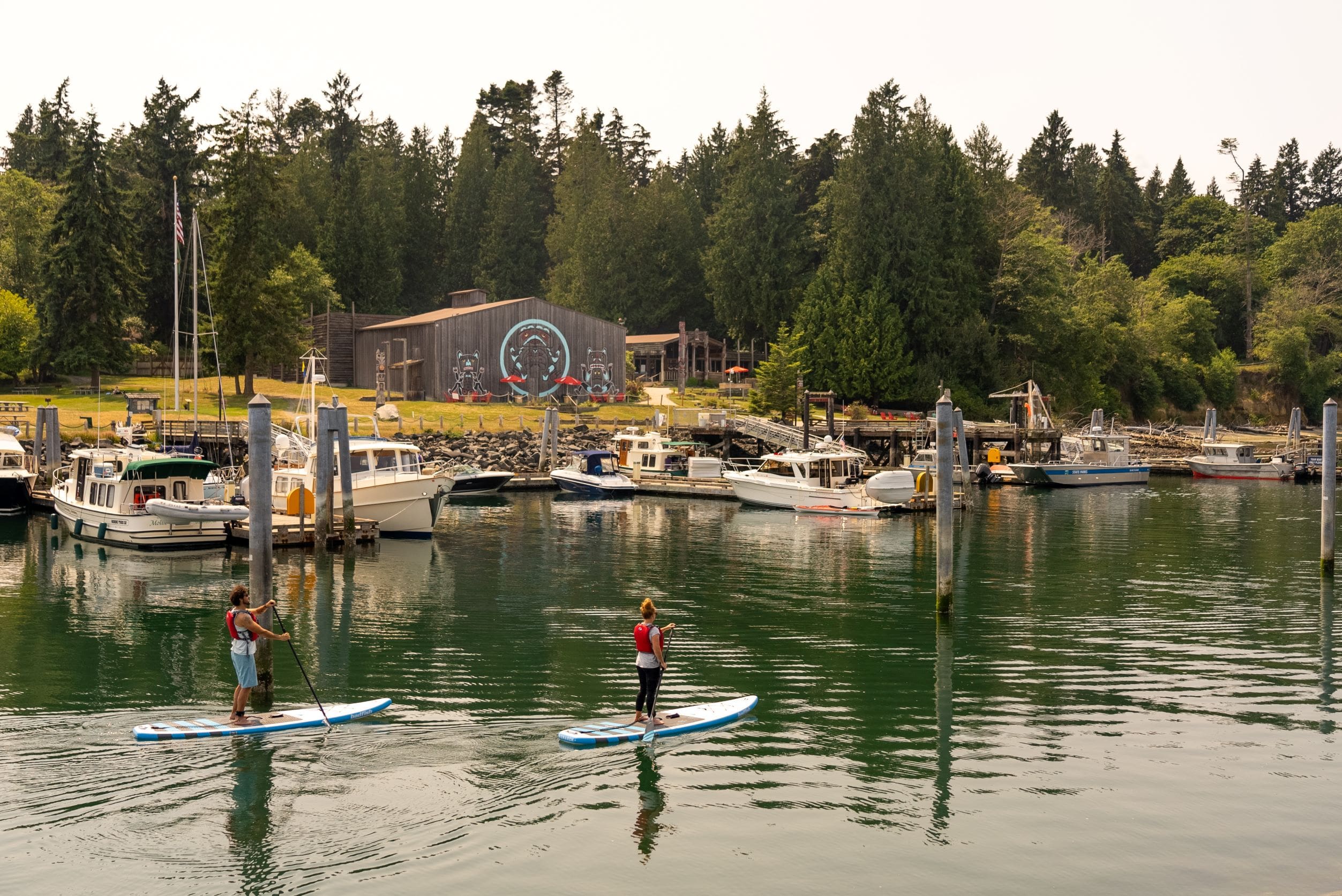 Man and woman paddle boarding in marina with boats in midground and longhouse in background with green trees surrounding it