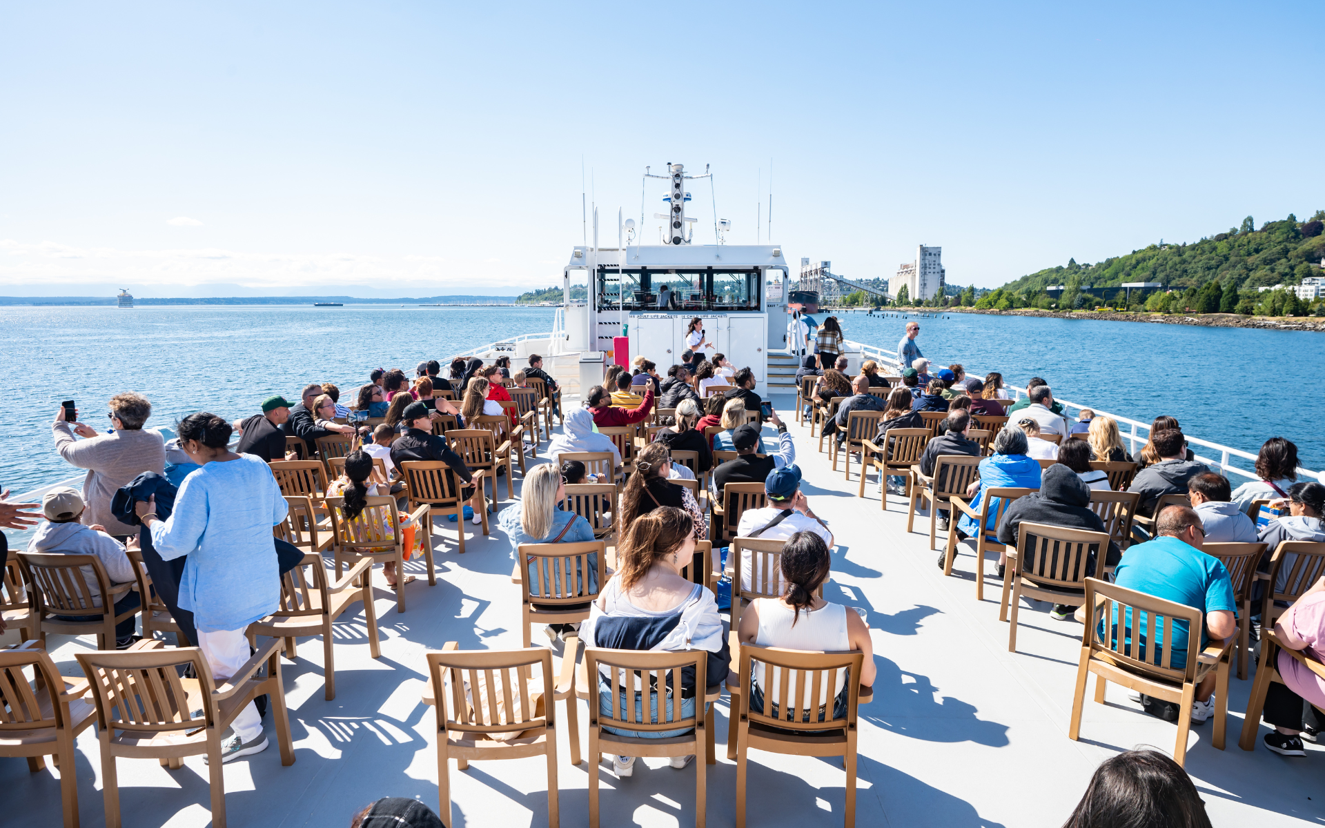 A diverse group of passengers enjoys a sunny boat ride, seated on deck. A city skyline and green hills line the water.