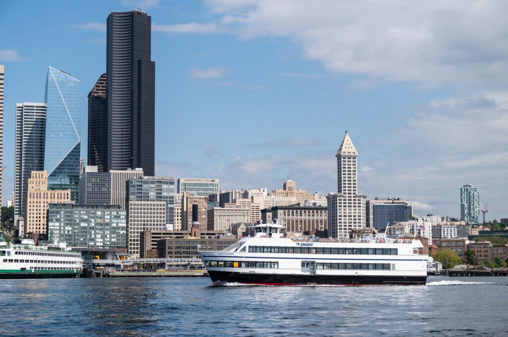 A white and black passenger ferry sails across water toward the right, with downtown Seattle's tall buildings under a blue sky.