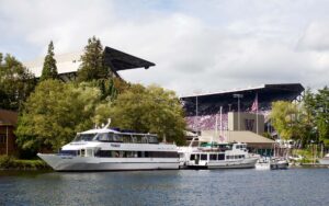 Boats docked on the water, with University of Washington's Husky Stadium and its purple-clad crowd in the background.