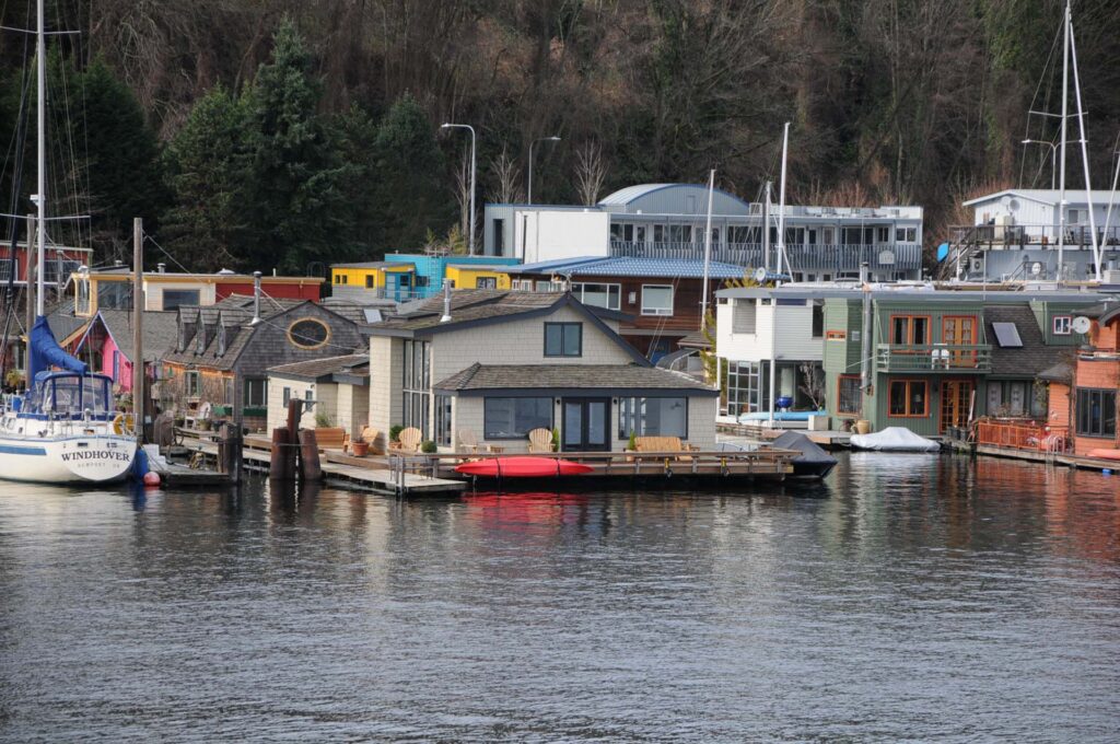 Vibrant houseboats and a sailboat line calm water, backed by a tree-covered hill.