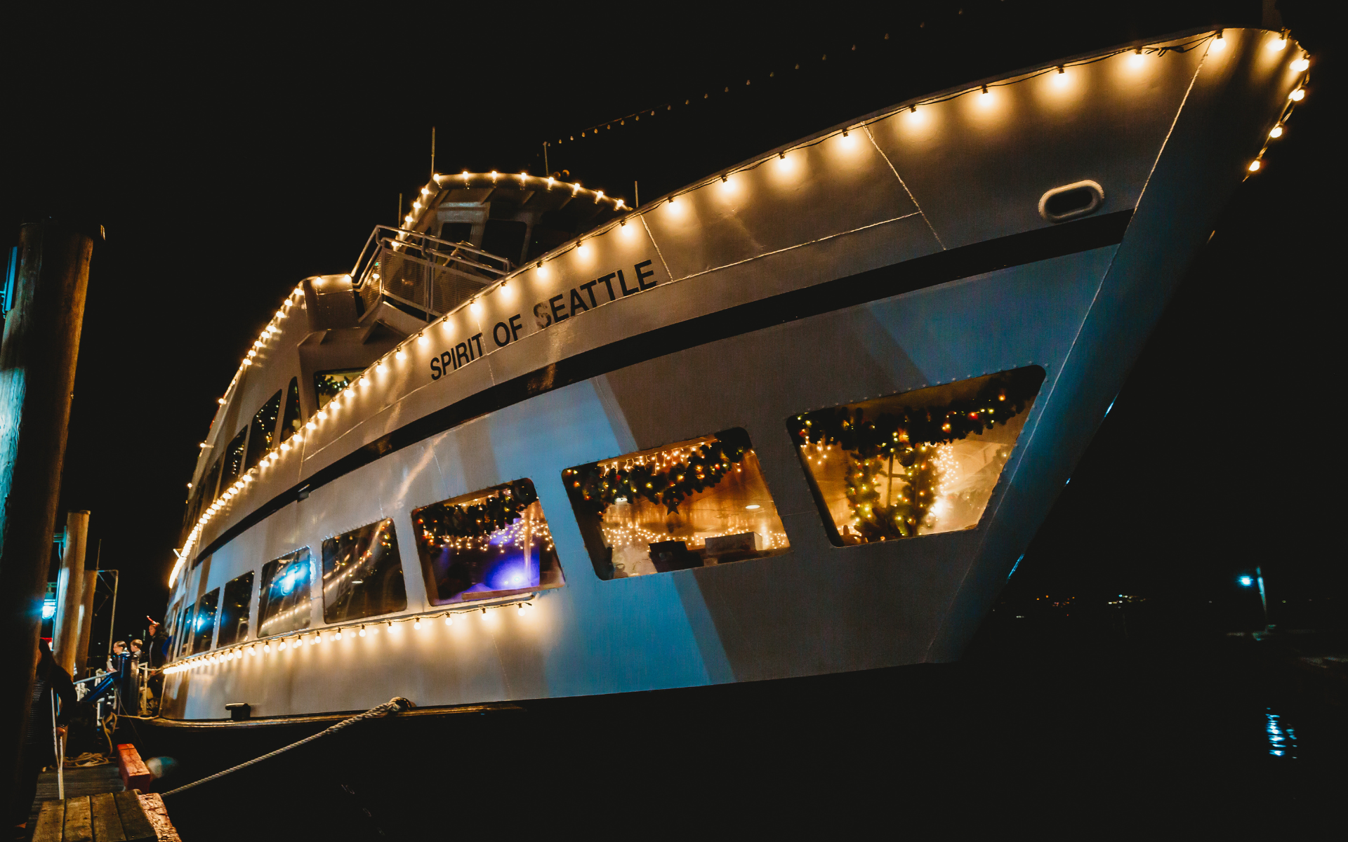 Spirit of Seattle boat sits docked at night, illuminated by rows of warm fairy lights along its multiple tiered decks.
