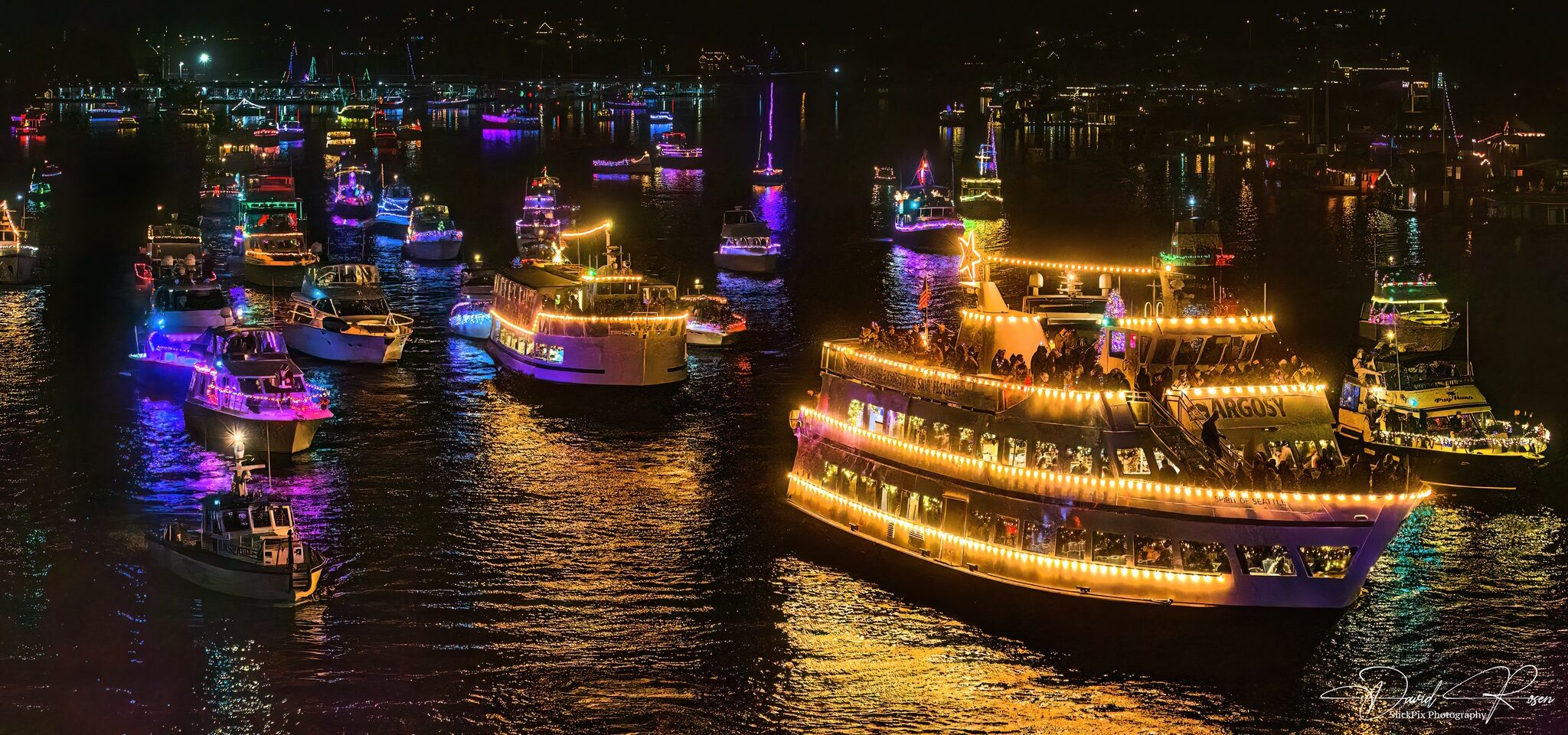 Christmas Ship Festival: Dozens of boats covered in dazzling Christmas lights cruise on dark water at night, part of a festive holiday ship parade.