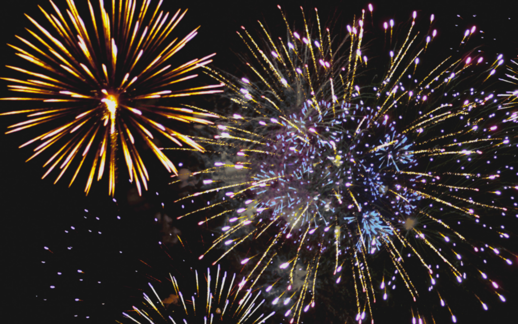 Multiple vibrant fireworks explode in the night sky, featuring golden starbursts and clusters of purple, blue, and white sparks.