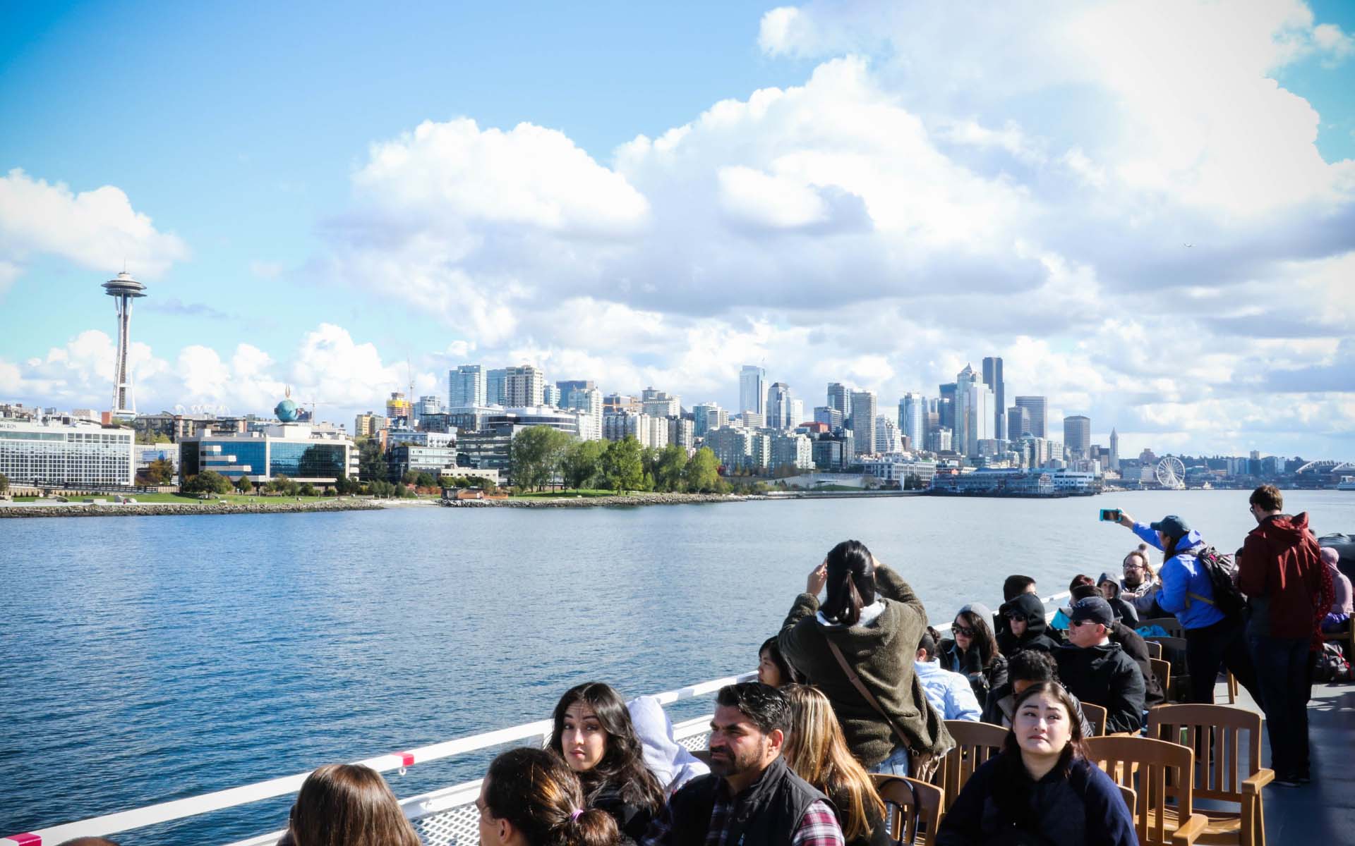 Tourists on a boat cruise enjoying views of the Seattle skyline, including the Space Needle, under a partly cloudy sky.