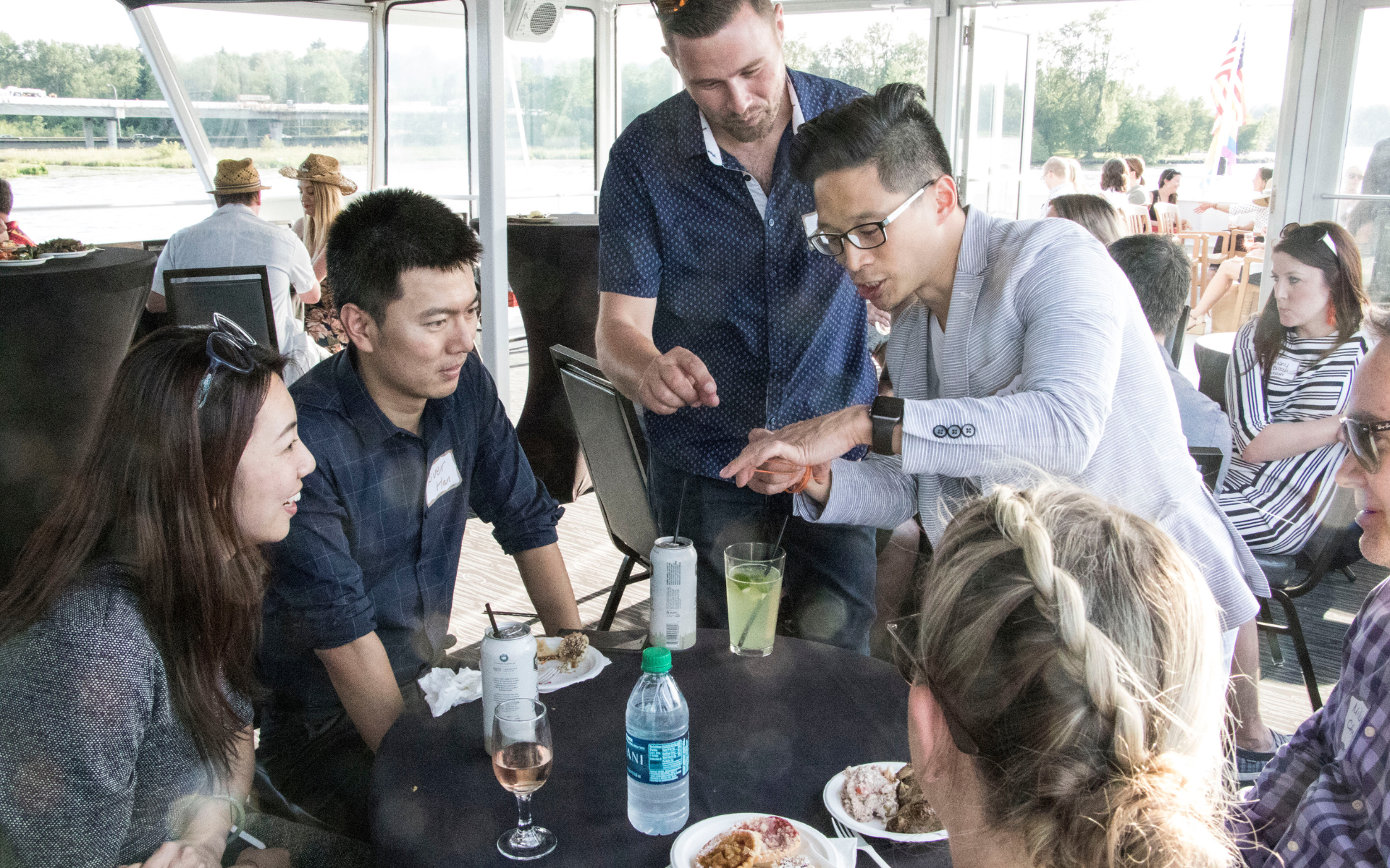 A diverse group of professionals enjoys a social event on a boat, with some gathered around a table watching an interaction.