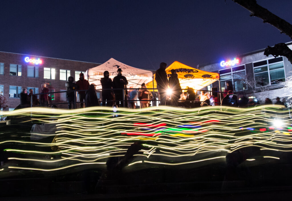 Silhouettes of people stand under white and yellow vendor tents at night, set against buildings with bright Google logos.