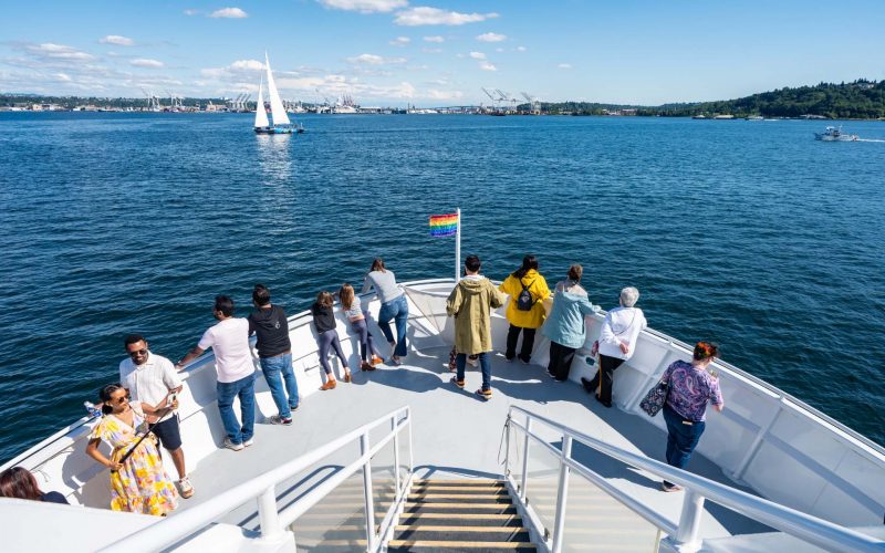 2026 Summer Soccer: A group enjoys a sunny boat cruise, looking towards a distant sailboat on the water. A rainbow flag flies at the bow.