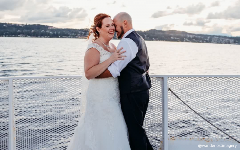 A joyful bride in a white gown and groom in a vest embrace on a ferry deck, laughing with a scenic water and mountain backdrop.