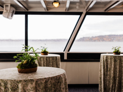 Three green-patterned tables with plant centerpieces by large windows, offering a wide view of water and distant land.