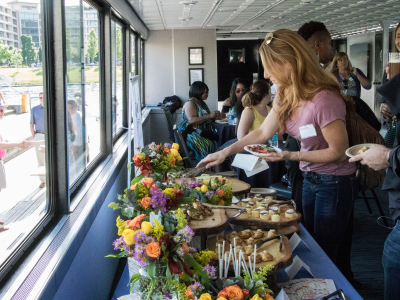 Attendees enjoy a social event on a boat, selecting food from a buffet adorned with flowers, while viewing a sunny outdoor waterfront scene.