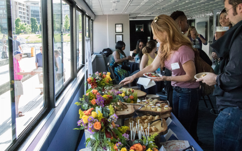 Attendees enjoy a social event on a boat, selecting food from a buffet adorned with flowers, while viewing a sunny outdoor waterfront scene.