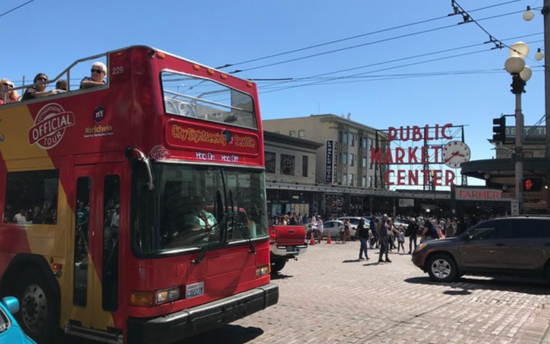 A red double-decker tour bus drives past Seattle's Public Market Center sign on a sunny day with pedestrians and cars.