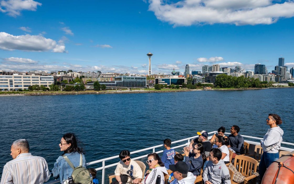 Harbor Cruise: Tourists on a boat view the Seattle skyline, including the Space Needle, while enjoying a sunny harbor cruise.