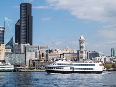 A white and black passenger ferry sails across water toward the right, with downtown Seattle's tall buildings under a blue sky.
