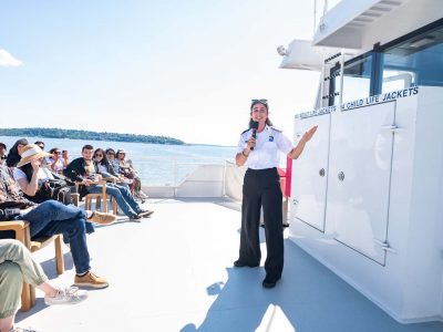 A guide addresses seated passengers on a sunny ferry deck. She holds a microphone, gesturing as the audience listens intently.