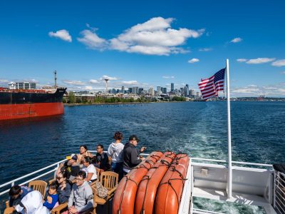 Boat travelers enjoy sunny views of the Seattle skyline, Space Needle, and a large cargo ship, with an American flag flying.