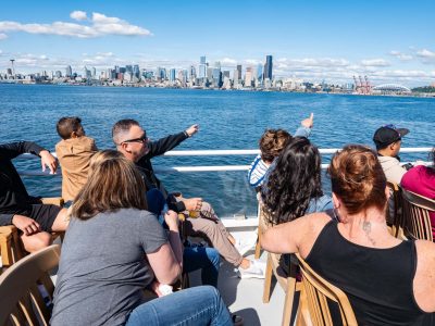 Passengers on a boat point towards the Seattle skyline with the Space Needle and other buildings, across blue water on a sunny day.