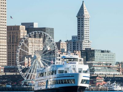 A ferry cruises Seattle's waterfront, passing the Great Wheel and historic Smith Tower against a clear blue sky.