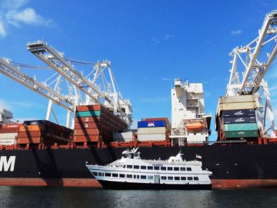 A small white passenger ferry sails alongside a massive black cargo ship loaded with colorful containers under towering port cranes.