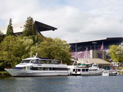 Boats docked on the water, with University of Washington's Husky Stadium and its purple-clad crowd in the background.