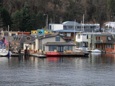 Vibrant houseboats and a sailboat line calm water, backed by a tree-covered hill.