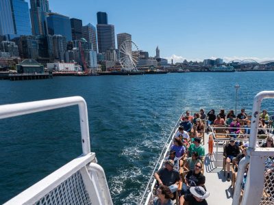 A boat full of people cruises through Puget Sound, passing Seattle's waterfront with its iconic Great Wheel and towering skyscrapers under a clear sky.