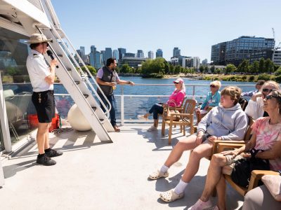 Passengers on a boat tour listen to a guide, smiling while cruising past a city waterfront on a clear day.