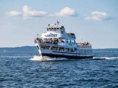 The Argosy Goodtime III ferry, packed with passengers on deck, cruises across calm blue water, leaving a white wake behind.