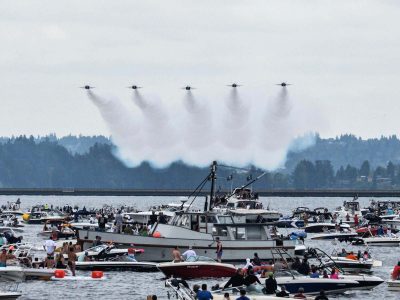 Seafair Blue Angels Viewing Cruise: Six Blue Angels jets perform a flyover, creating white contrails above a water body bustling with numerous spectator boats.