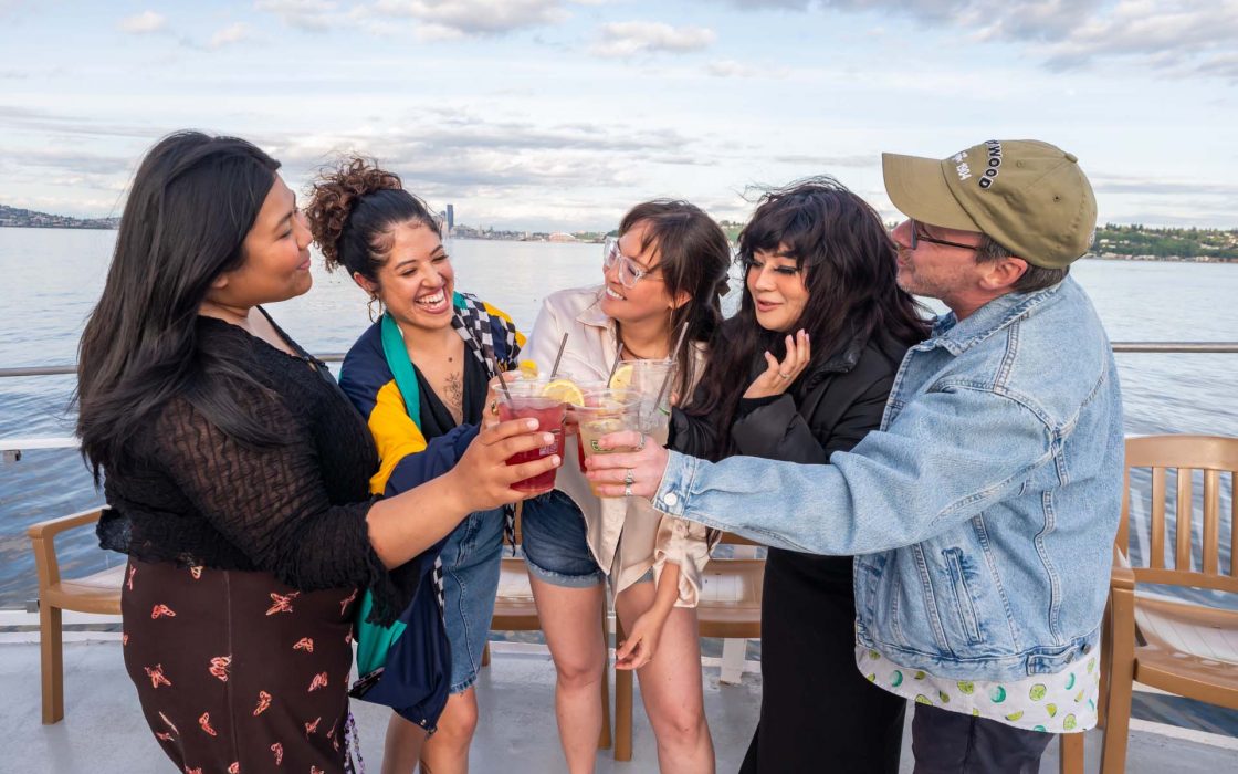 Seattle Happy Hour Cruise: Diverse group of five celebrating on a boat, toasting drinks with smiles against a scenic Seattle waterfront backdrop.
