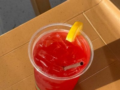 Refreshing red beverage with ice, a lemon garnish, and a straw in a clear cup, placed on a textured tan surface under bright sun.