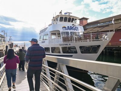 People walk on a pier gangway toward the water, where the Argosy Goodtime III tour boat is docked under a partly cloudy sky.