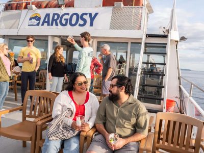 Couples and friends enjoying drinks on an Argosy ferry cruise under a blue sky. The man and woman in front smile at each other.