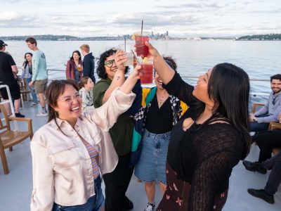 Four joyful women making a toast with colorful drinks on a boat. Other guests socialize with the Seattle skyline in the distance.