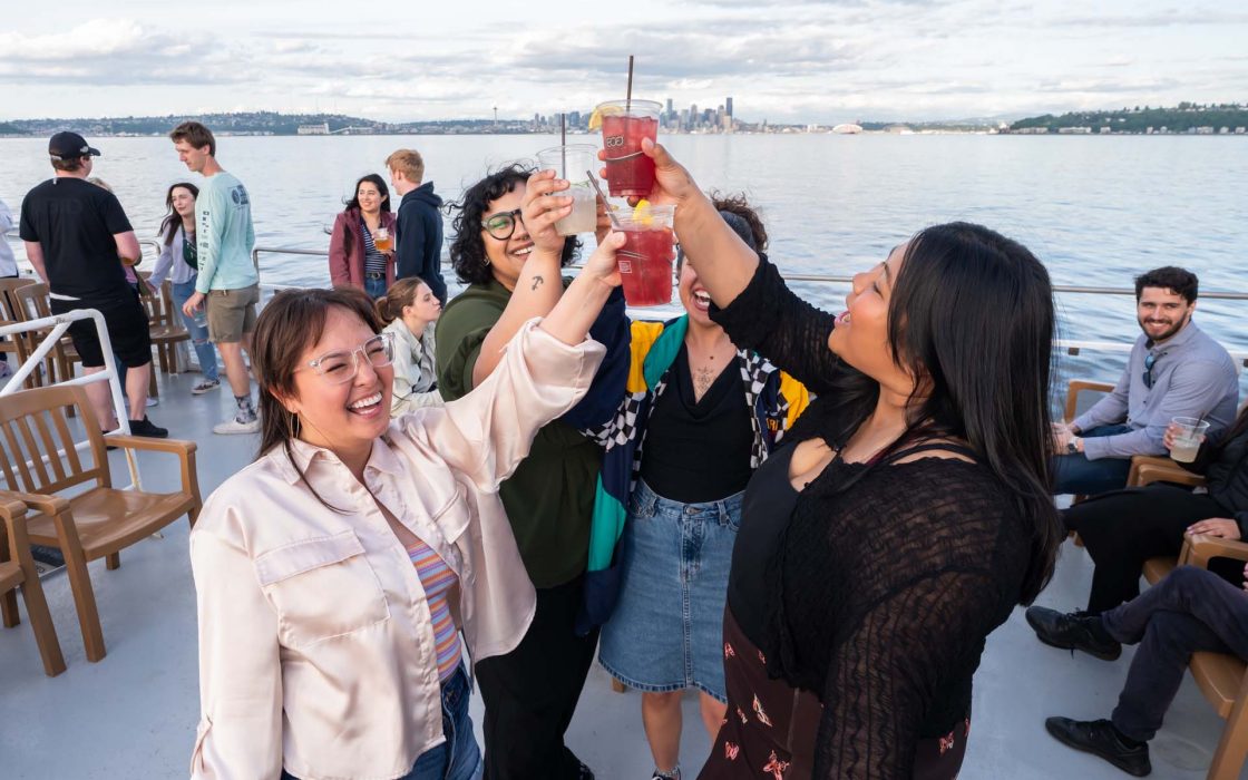 Four joyful women making a toast with colorful drinks on a boat. Other guests socialize with the Seattle skyline in the distance.