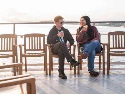Two people, a man in sunglasses and a woman, smiling and holding drinks while sitting on chairs on a boat deck in the sun.