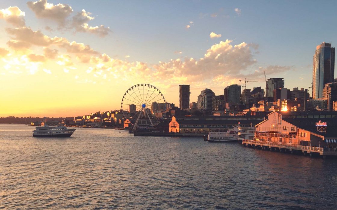 Seattle Sunset Cruise: Seattle waterfront at sunset with a golden sky, showing the Great Wheel, downtown skyline, and boats on the water.