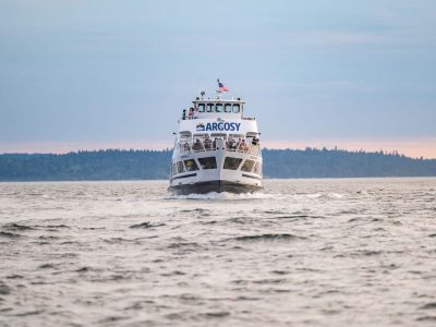 A white multi-deck Argosy boat, full of passengers, sails forward on the water. A dark forested shoreline lies under a soft evening sky.