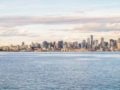 Seattle cityscape across calm water, featuring skyscrapers, the Space Needle, and a faint airplane in the cloudy sky.